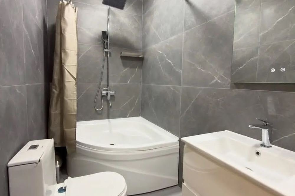 A grey tiled bathroom with a corner bath and a lit mirror